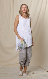 Front full body view of a woman wearing the chalet lucianna tunic. This tunix is sleeveless. It is white with multicolored patchwork trim and a front draped multicolored patchwork pocket. The tunic has an asymmetrical hem. On the bottom the model is wearing grey capris.