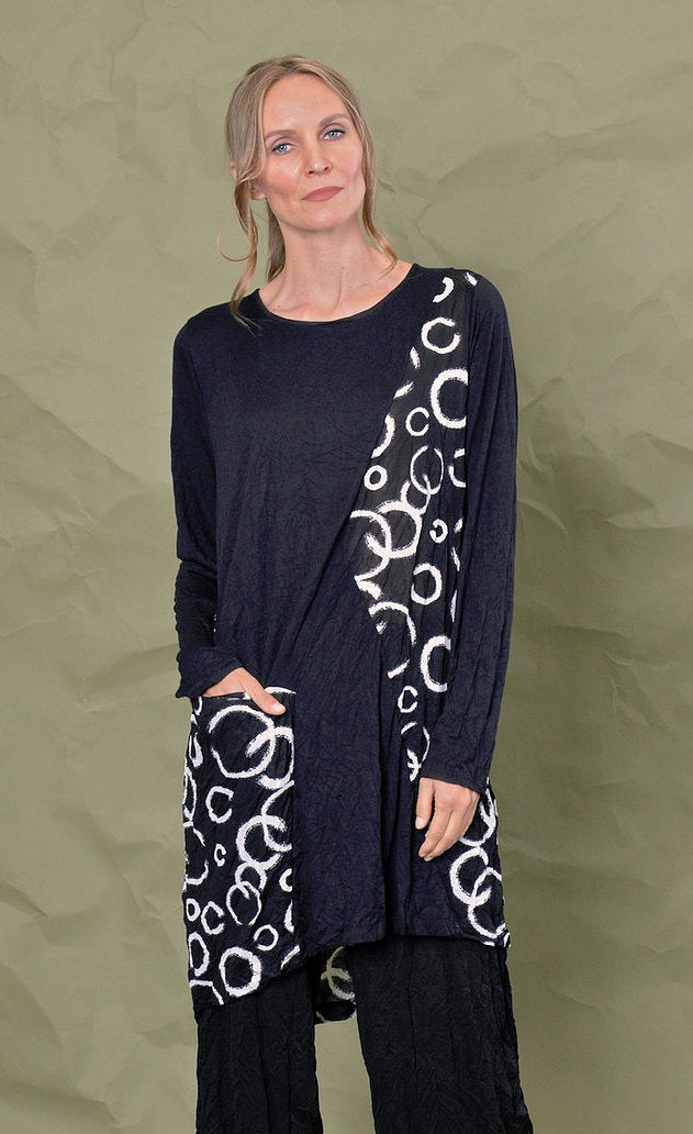 Front top half view of a woman wearing black pants and the chalet onyx pippa tunic. This black tunic has long sleeves, patches of white circle print and a pocket with the same print on it. The model has her hand in the front pocket.