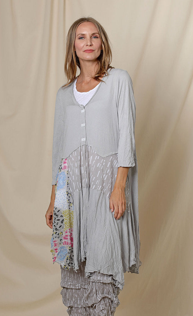 Front top half view of a woman wearing the chalet orla jacket. The jacket in this image is grey with a patchwork print on the right side, a 3 button front, 3/4 length sleeves, and an asymmetrical hem.