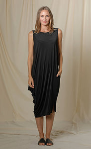 Front full body view of a woman wearing the chalet romaine dress. This dress is black and sleeveless. The right side has a draping effect and the dress sits slightly below the knees.