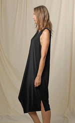 Load image into Gallery viewer, Left side full body view of a woman wearing the chalet romaine dress. This dress is black and sleeveless. The right side has a draping effect and the dress sits slightly below the knees.
