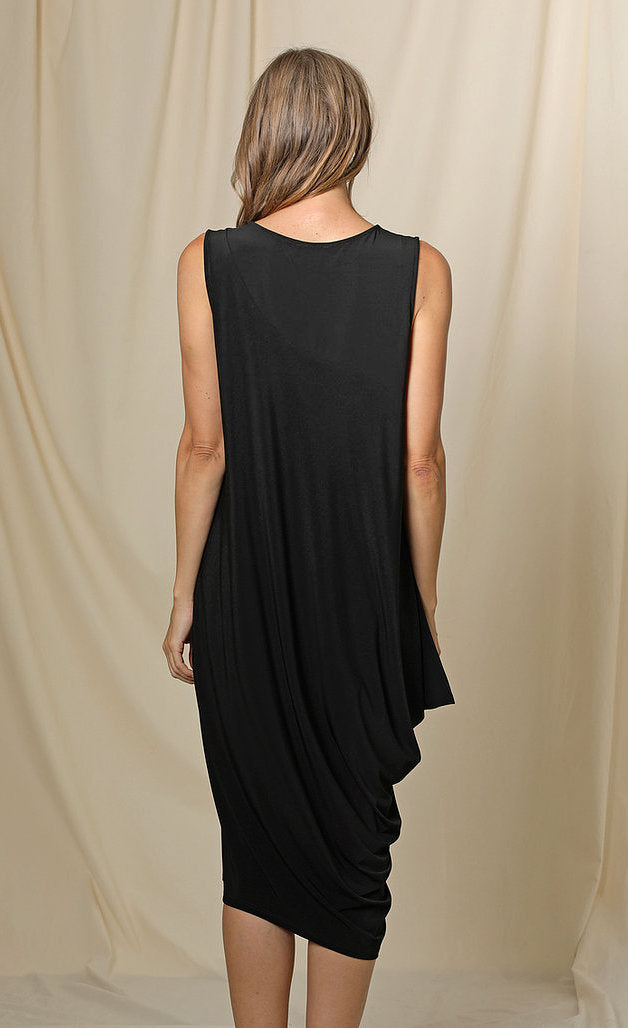 Back full body view of a woman wearing the chalet romaine dress. This dress is black and sleeveless. The right side has a draping effect and the dress sits slightly below the knees.