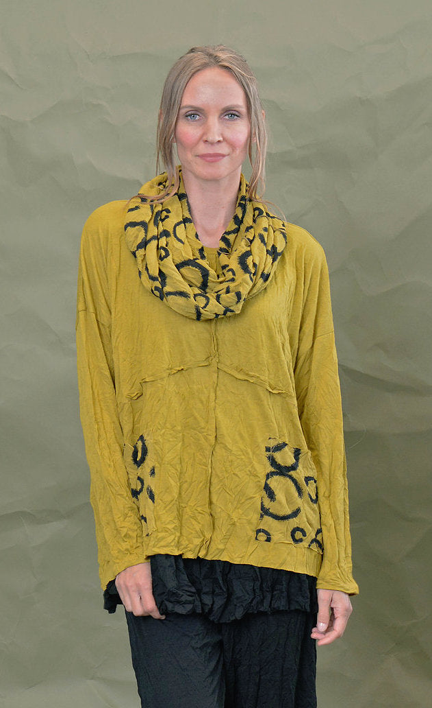 Front top half view of a woman wearing the Chalet Simone Top in the color marigold. This top has long sleeves, decorative criss-crossing seams and two front pockets with black circles on them.