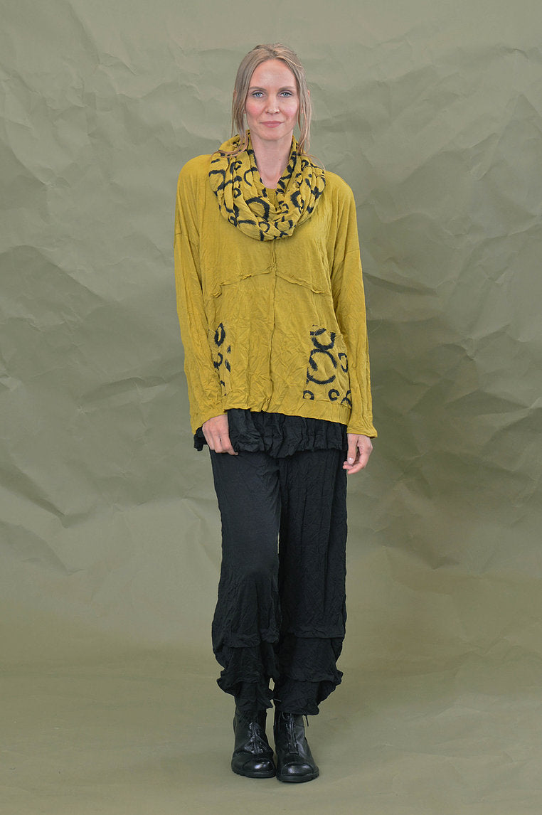 Front full body view of a woman wearing black pants and the Chalet Simone Top in the color marigold. This top has long sleeves, decorative criss-crossing seams and two front pockets with black circles on them.