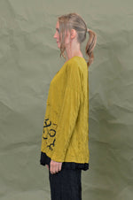 Load image into Gallery viewer, Left side top half view of a woman wearing the Chalet Simone Top in the color marigold. This top has long sleeves, decorative criss-crossing seams and two front pockets with black circles on them.

