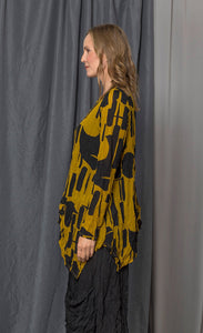 Side top half view of a woman wearing the chalet thora top. This top is gold and black with a geometric print. The top has long sleeves and an asymmetrical pointed hem.