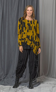 Front full body view of a woman wearing the chalet thora top. This top is gold and black with a geometric print. The top has long sleeves and an asymmetrical pointed hem.