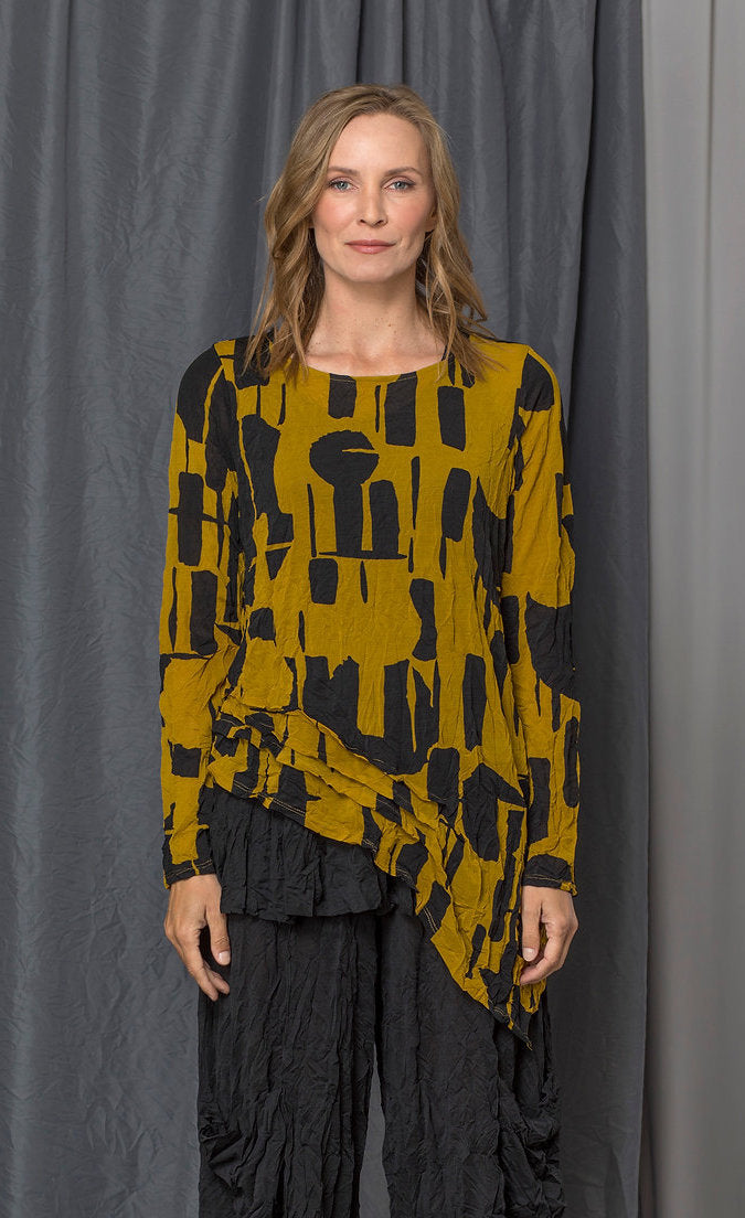 Front top half view of a woman wearing the chalet thora top. This top is gold and black with a geometric print. The top has long sleeves and an asymmetrical pointed hem.