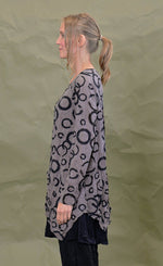 Load image into Gallery viewer, Left side top half view of a woman wearing the chalet cinder thora top. This top is grey with black circles. It has long sleeves and an asymmetrical hem with a long left side in the front.

