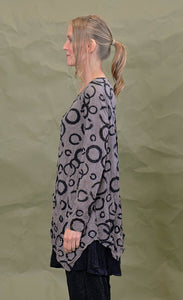 Left side top half view of a woman wearing the chalet cinder thora top. This top is grey with black circles. It has long sleeves and an asymmetrical hem with a long left side in the front.