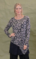 Load image into Gallery viewer, Front top half view of a woman wearing the chalet cinder thora top. This top is grey with black circles. It has long sleeves and an asymmetrical hem with a long left side in the front.
