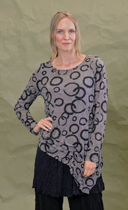 Front top half view of a woman wearing the chalet cinder thora top. This top is grey with black circles. It has long sleeves and an asymmetrical hem with a long left side in the front.