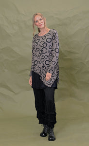 Front full body view of a woman wearing the chalet cinder thora top. This top is grey with black circles. It has long sleeves and an asymmetrical hem with a long left side in the front.
