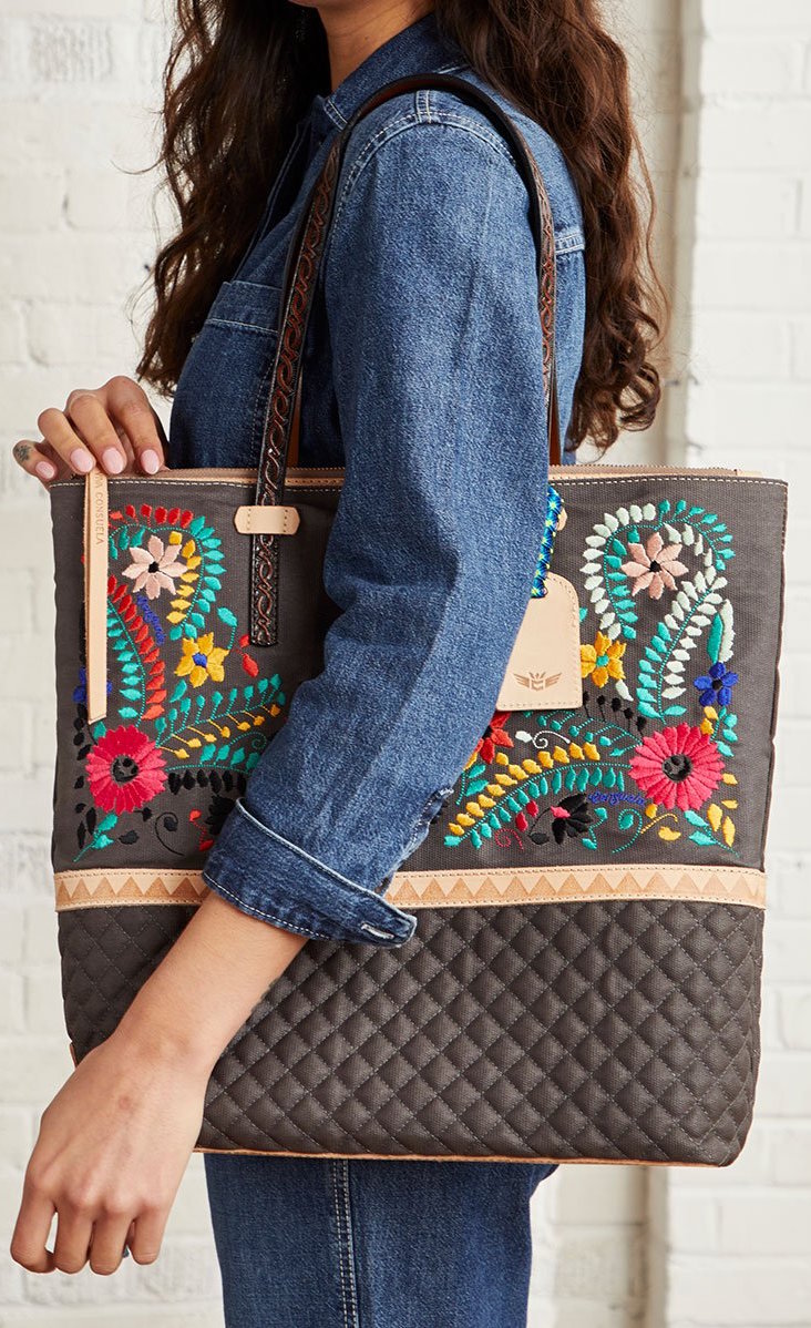 Front view of a model carrying the consuela silverlake market tote on her shoulder. This tote is grey with a quilted like bottom and light tan trim. The straps are thin. On the front of the tote is blue pink and orange floral embroidery.