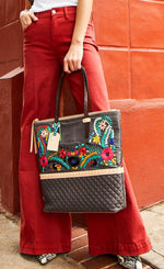 Load image into Gallery viewer, Front view of a model in red pants leaning on a red building and carrying the consuela silverlake market tote. This tote is grey with a quilted like bottom and light tan trim. The straps are thin. On the front of the tote is blue pink and orange floral embroidery.
