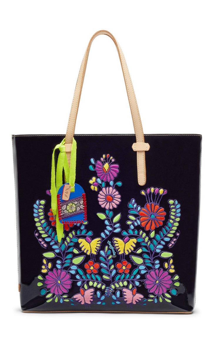 Front view of the consuela tia slim tote. This tote is a glossy black with a thin tan leather strap and colorful embroidery on the front.