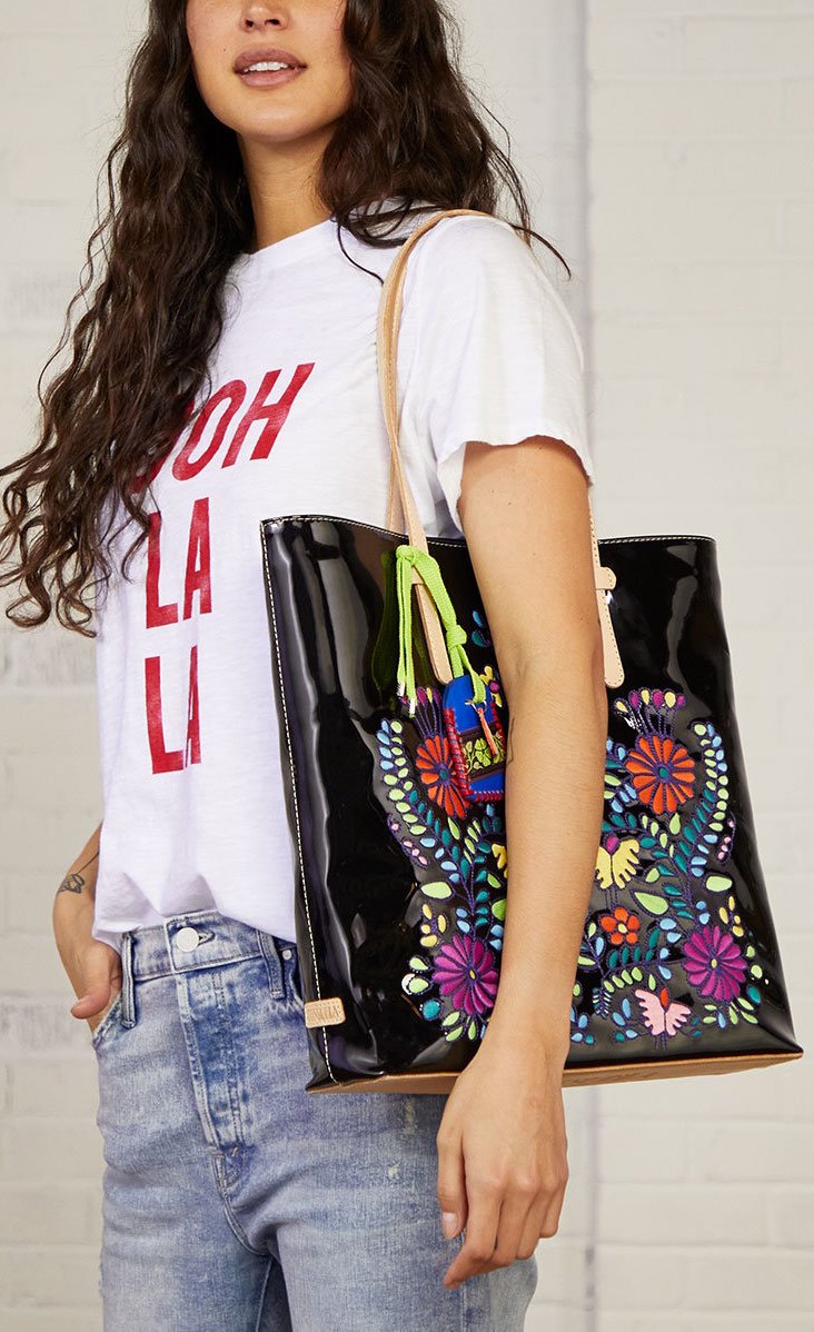 Front view of a woman carrying the consuela tia slim tote. This tote is a glossy black with a thin tan leather strap and colorful embroidery on the front.