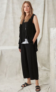 Front full body view of a woman wearing the crea concept black vest/top. This top is sleeveless with a draped front, a zipper on the front left side, and an asymmetrical hem.
