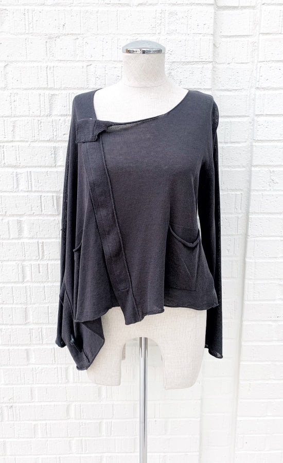 Front view of the crea concept knit cape top. This top is grey and semi-translucent. It has long sleeves, two patch front pockets, a right sided snap up front, and a cape feature over the right arm.