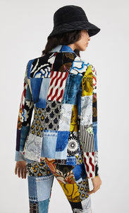 Back top half view of a woman wearing a hat and the desigual alejandra denim patch blazer. This blazer features a patchwork of multiple prints, a single button closure, a flat lapel, and long sleeves.