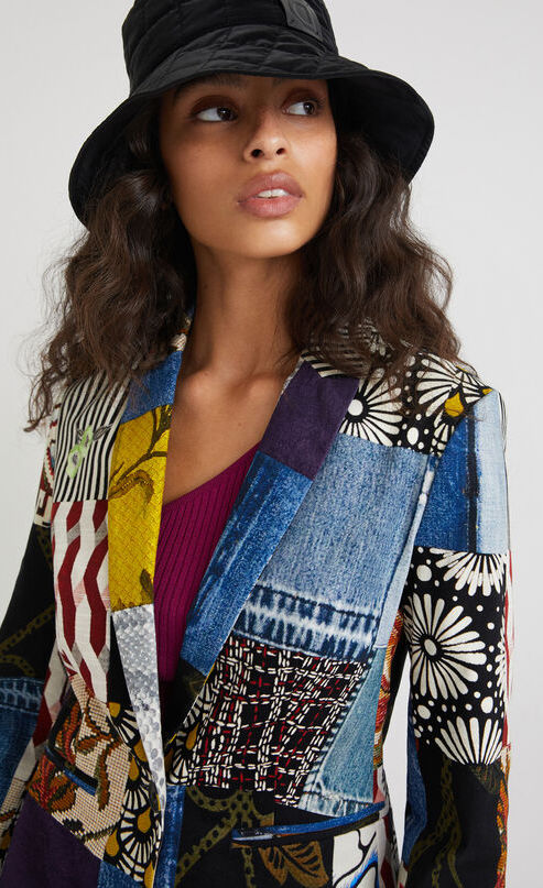 Front top half view of a woman wearing a hat and the desigual alejandra denim patch blazer. This blazer features a patchwork of multiple prints, a single button closure, a flat lapel, and long sleeves.