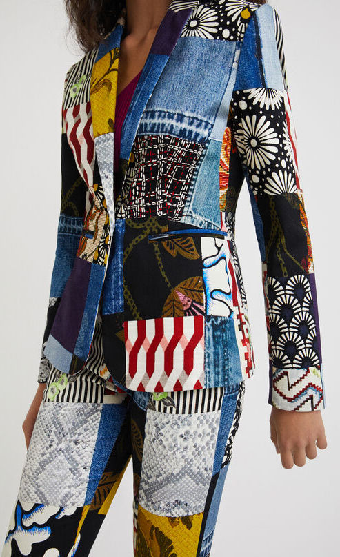 Front close up view of a woman wearing a hat and the desigual alejandra denim patch blazer. This blazer features a patchwork of multiple prints, a single button closure, a flat lapel, and long sleeves.
