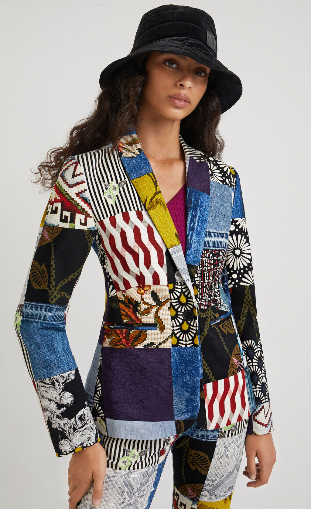 Front top half view of a woman wearing a hat and the desigual alejandra denim patch blazer. This blazer features a patchwork of multiple prints, a single button closure, a flat lapel, and long sleeves.