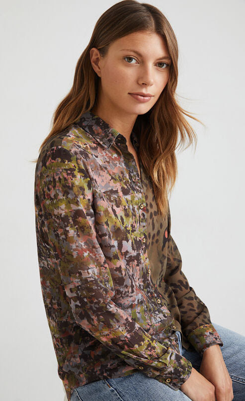 Front top half view of a woman wearing the desigual daytona wild print shirt. The base color of this shirt is green/brown. It has an animal print on the left side and a mixed watercolor print on the right side. The shirt has a button up front and long sleeves with cuffs.