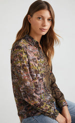 Load image into Gallery viewer, Front top half view of a woman wearing the desigual daytona wild print shirt. The base color of this shirt is green/brown. It has an animal print on the left side and a mixed watercolor print on the right side. The shirt has a button up front and long sleeves with cuffs.
