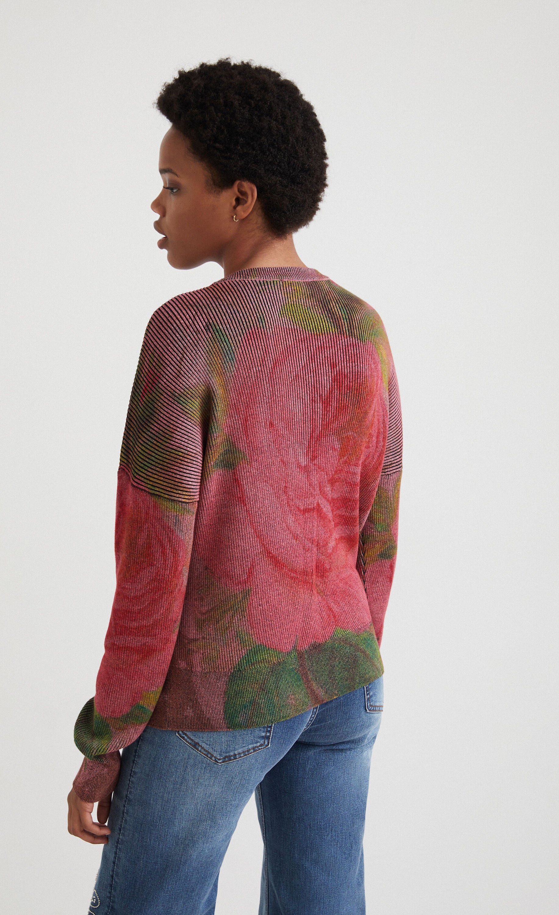 back top half view of a woman wearing the desigual knit flowers sweater. This sweater is ribbed with large pink roses printed all over it. It has long sleeves and a loose fit.