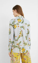 Load image into Gallery viewer, Back top half view of a woman wearing yellow paisley capris and the desigual lemons cotton shirt. This shirt is blue with lemons printed on it. The back of the top is yoked and has an inverted pleat. The sleeves are long.
