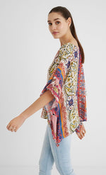 Load image into Gallery viewer, Left side top half view of a woman wearing a the desigual paisley blouse. This blouse looks like a poncho with arm holes. It has a multicolored, pink dominate paisley pattern on it. 
