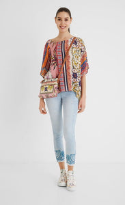 Front full body view of a woman wearing a the desigual paisley blouse. This blouse looks like a poncho with arm holes. It has a multicolored, pink dominate paisley pattern on it. 