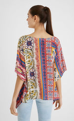 Load image into Gallery viewer, Back top half view of a woman wearing a the desigual paisley blouse. This blouse looks like a poncho with arm holes. It has a multicolored, pink dominate paisley pattern on it. 
