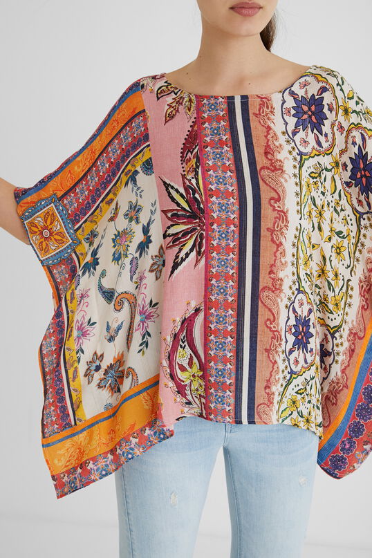 Front top half view of a woman with her arms up and wearing a the desigual paisley blouse. This blouse looks like a poncho with arm holes. It has a multicolored, pink dominate paisley pattern on it. 