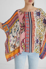 Load image into Gallery viewer, Front top half view of a woman with her arms up and wearing a the desigual paisley blouse. This blouse looks like a poncho with arm holes. It has a multicolored, pink dominate paisley pattern on it. 
