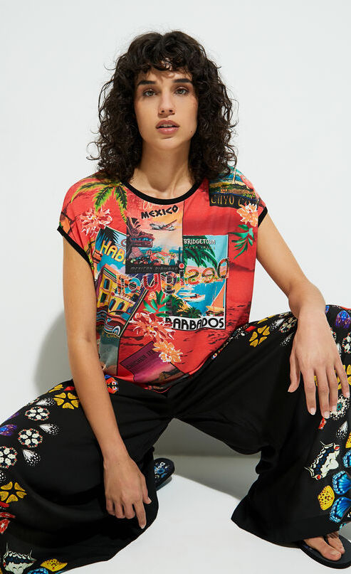 Front full body view of a woman crouched down and wearing the desigual postcards short sleeve t-shirt. This top is red with tropical post cards all over it and flowers. The top has short sleeves, a round neck, and a rounded hem.