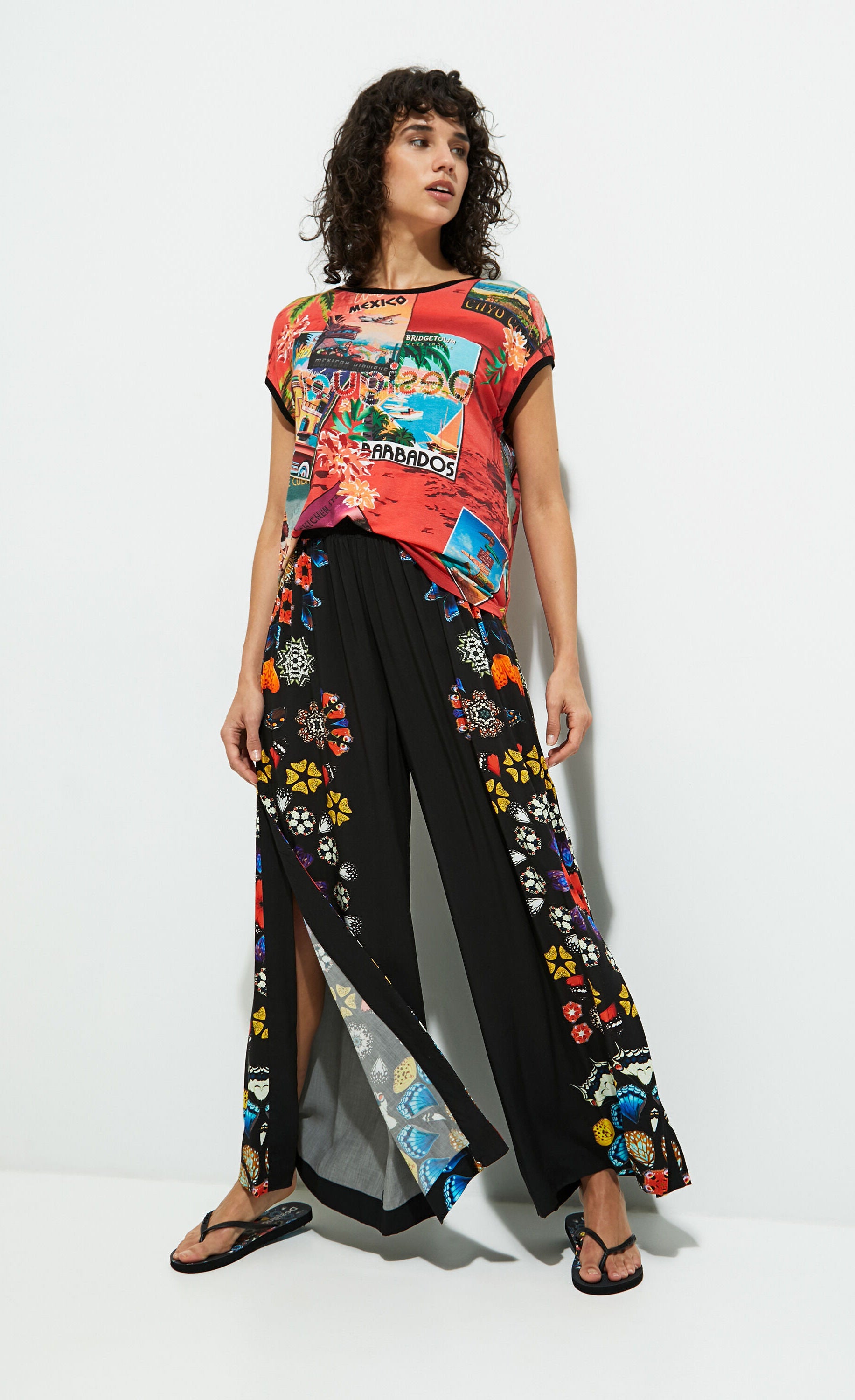 Front full body view of a woman wearing the desigual postcards short sleeve t-shirt. This top is red with tropical post cards all over it and flowers. The top has short sleeves, a round neck, and a rounded hem.