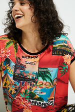 Load image into Gallery viewer, Front close up view of a woman wearing the desigual postcards short sleeve t-shirt. This top is red with tropical post cards all over it and flowers. The top has short sleeves and a round neck.
