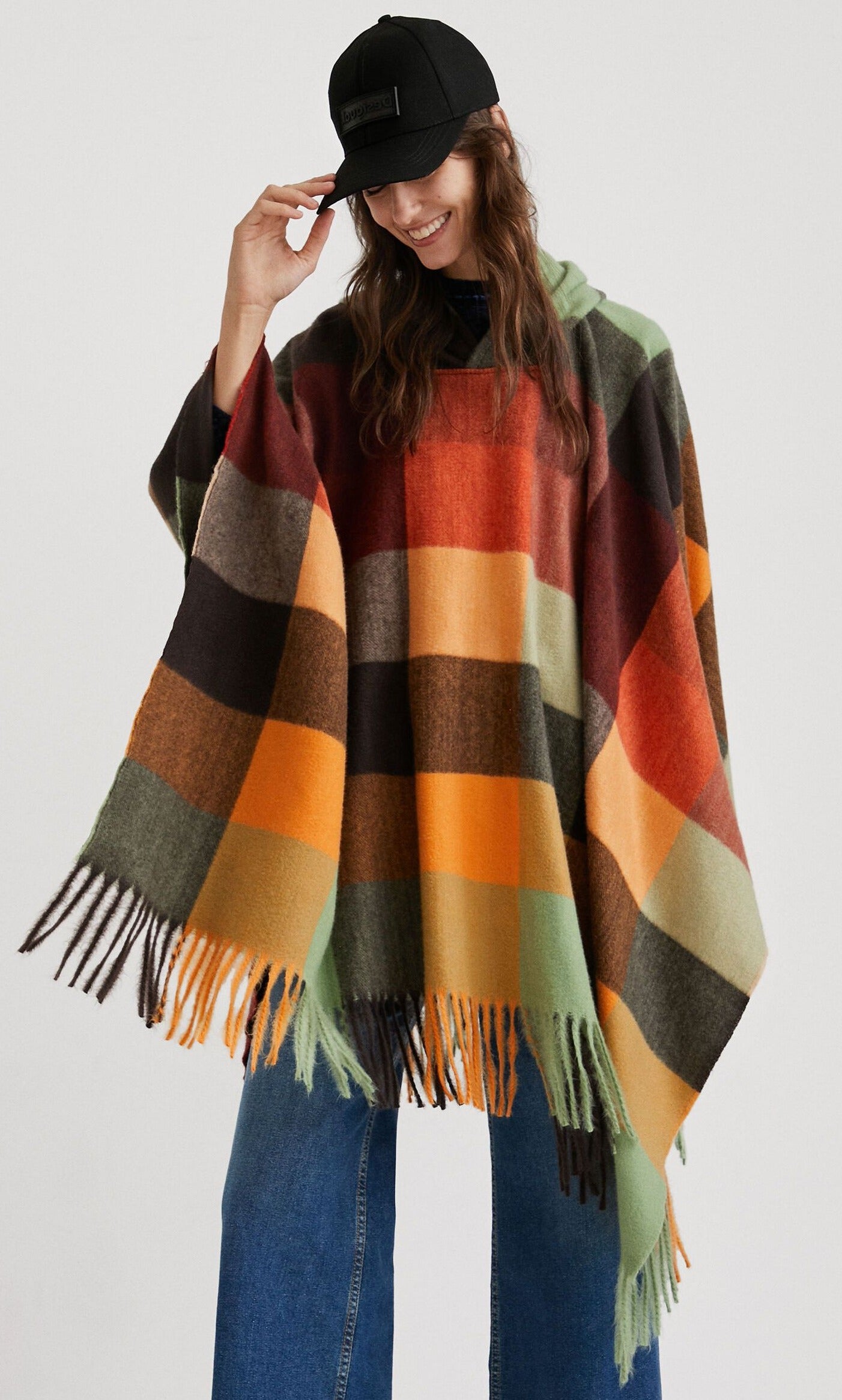 Front top half view of a woman wearing the desigual plaid poncho with wide jeans. This poncho is brown, red, orange, and green plaid. It has a hood and a fringed hem.