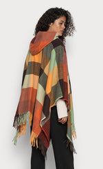 Load image into Gallery viewer, Back top half view of a woman wearing the desigual plaid poncho over a white top with black pants. This poncho is brown, red, orange, and green plaid. It has a hood and a fringed hem.
