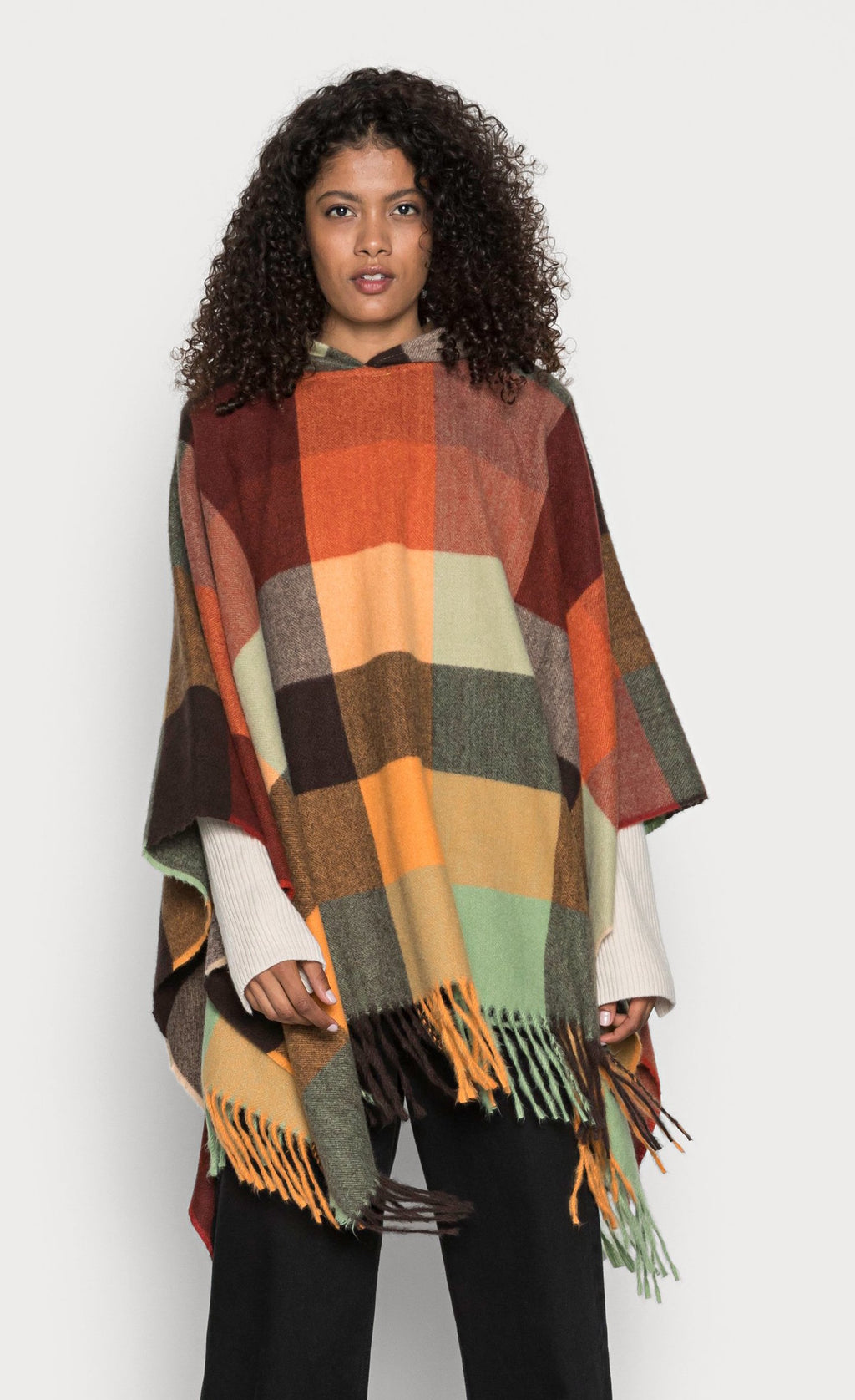 Front top half view of a woman wearing the desigual plaid poncho over a white top with black pants. This poncho is brown, red, orange, and green plaid. It has a hood and a fringed hem.