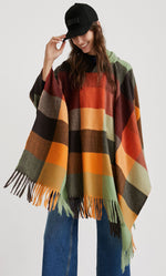 Load image into Gallery viewer, Front top half view of a woman wearing the desigual plaid poncho with wide jeans. This poncho is brown, red, orange, and green plaid. It has a hood and a fringed hem.
