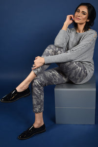 Front full body view of a woman sitting and wearing the frank lyman grey knit sweater. This solid sweater has long sleeves with a braided detail running down the sides. 
