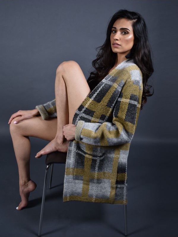 Left side full body view of a woman sitting on a chair and wearing the frank lyman plaid sweater jacket. This jacket is grey and mustard with long sleeves and two front pockets. 