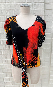 Front view of a mannequin wearing the black and orange tie top from frank lyman. This top has a print that features orange paint stroke-like shapes and white dots on a black fabric. The top has ruffled sleeves that end at the elbow and a front, center tie at the bottom.