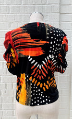 Load image into Gallery viewer, Back view of a mannequin wearing the black and orange tie top from frank lyman. This top has a print that features orange paint stroke-like shapes and white dots on a black fabric. The top has ruffled sleeves that end at the elbow.
