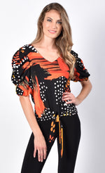 Load image into Gallery viewer, Front view of a woman with her left hand on her hip wearing black pants and a black and orange tie top from frank lyman. This top has a print that features orange paint stroke-like shapes and white dots on a black fabric. The top has ruffled sleeves that end at the elbow and a front, center tie at the bottom.
