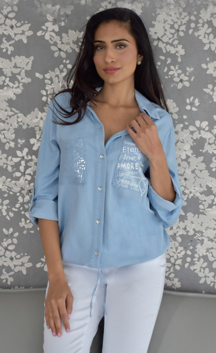 Front top half view of a woman wearing white pants and the frank lyman blue amore shirt. This shirt has a button down front, a drawstring hem with a tie in the middle, and two front patch pockets. The right pocket has sequins on it while the left pocket has words about love written on it.