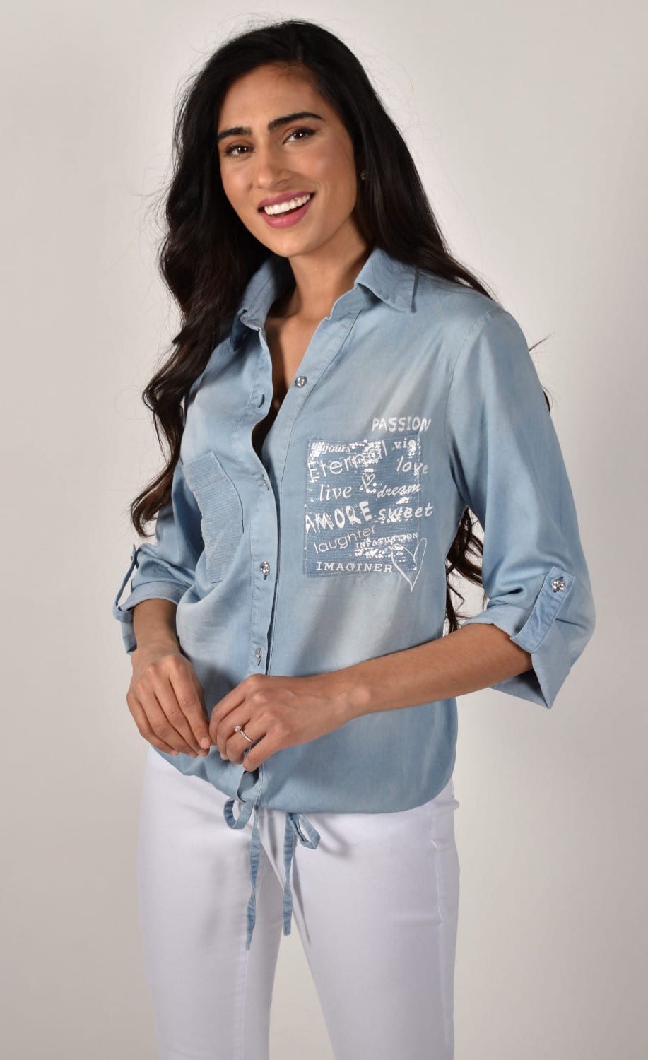 Front close up view of a woman wearing white pants and the frank lyman blue amore shirt. This shirt has a button down front, a drawstring hem with a tie in the middle, and two front patch pockets. The right pocket has sequins on it while the left pocket has words about love written on it.